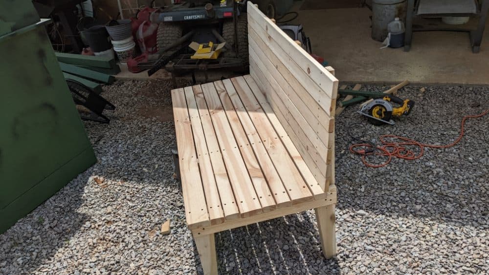 Wooden bench made of slatted planks, set on a gravel surface with tools in background.